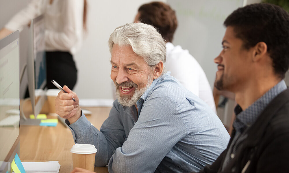 an older man and a younger man sit at a desk together working on their computer, dispelling generational differences